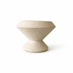 May include: A cream-colored ceramic bowl with a distinct hourglass shape, featuring a wide, angled rim that tapers down to a narrow center before widening again at the base. The surface has a rough, textured, and speckled finish throughout.