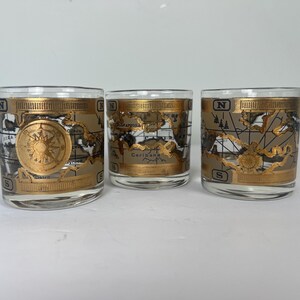 May include: Three clear drinking glasses with a gold map design. Each glass features a gold compass rose and a map of the world. The glasses have a gold rim and are labeled with the letters N and S.