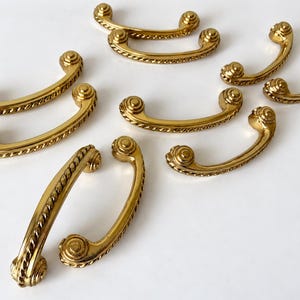 May include: A collection of eight gold-toned metal drawer pulls, each featuring a curved handle with a twisted rope texture along the center and circular, ridged end caps. The hardware is arranged in a scattered pattern across the frame.