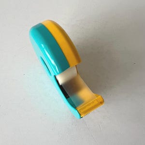 May include: A teal and yellow tape dispenser with a roll of clear tape. The dispenser has a rounded top and a cutting edge. The tape is partially dispensed, showing the adhesive side. The dispenser is made of plastic.