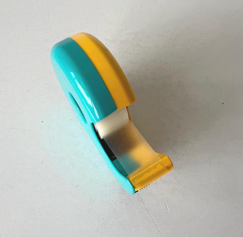 May include: A teal and yellow tape dispenser with a roll of clear tape. The dispenser has a rounded top and a cutting edge. The tape is partially dispensed, showing the adhesive side. The dispenser is made of plastic.