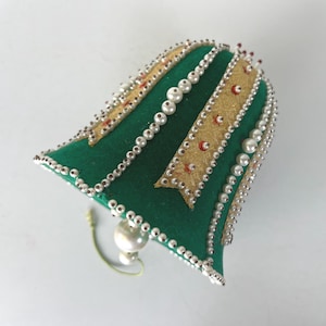 May include: A green bell-shaped ornament with gold ribbon accents and white pearl embellishments. The ornament has a white pearl at the bottom and a green string for hanging. The bell is likely for holiday or seasonal decor.