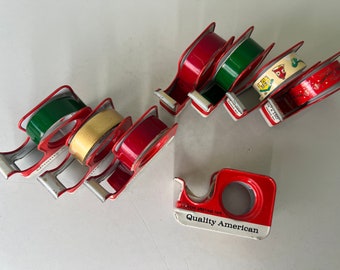 May include: A collection of vintage red, green, and gold tape dispensers with various designs. The dispensers are made of metal and have a retro look. One dispenser has the text "Quality American" printed on it.