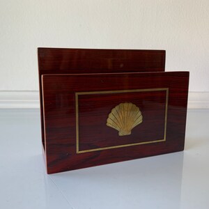 May include: A dark red wooden letter holder with a gold seashell design. The holder has a rectangular shape and a slightly raised edge.