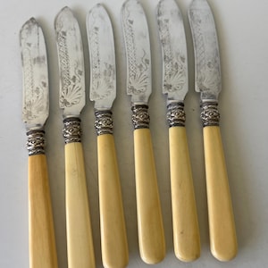May include: Set of six antique butter knives. Each knife features a silver blade with etched floral designs and a cream-colored handle. The handles are connected to the blades with a decorative silver collar. The knives are arranged in a row.
