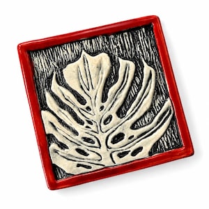 May include: A square ceramic tile featuring a raised, off-white monstera leaf design centered on a textured black background. The tile is framed by a glossy, vibrant red border, creating a distinct contrast between the organic leaf shape and the geometric red edge.