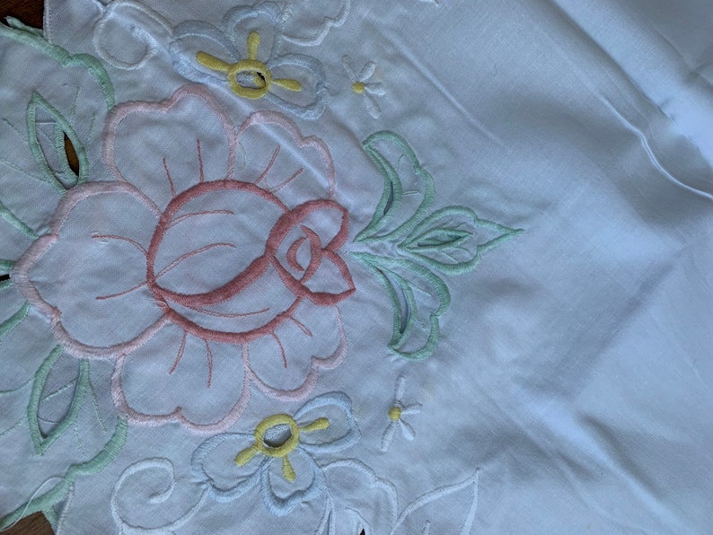 May include: White cotton fabric with embroidered flowers in pink, green, blue, and yellow. The flowers are in a floral pattern.