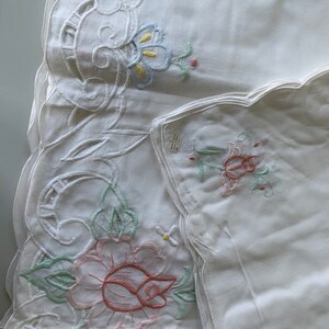 May include: Two white embroidered tablecloths with scalloped edges. One tablecloth features a large pink rose with green leaves and a smaller white flower. The other tablecloth has a smaller floral design with blue, yellow, and green accents.