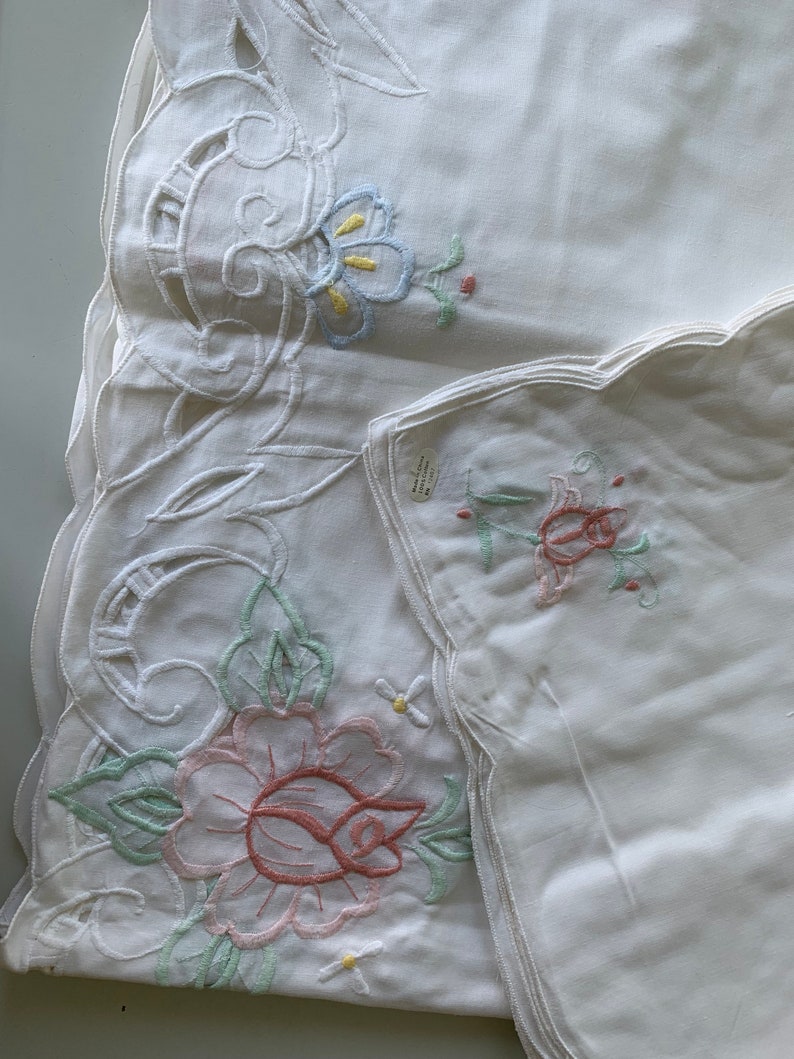 May include: Two white embroidered tablecloths with scalloped edges. One tablecloth features a large pink rose with green leaves and a smaller white flower. The other tablecloth has a smaller floral design with blue, yellow, and green accents.