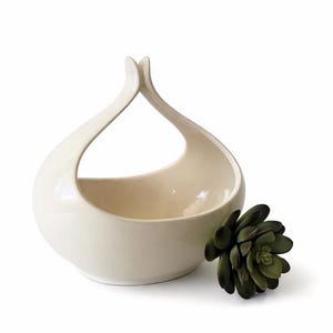 May include: A cream-colored ceramic bowl with a unique handle design. The bowl has a rounded base and a wide opening, with a decorative succulent plant to the side. The handle curves upwards, creating an elegant, modern look.