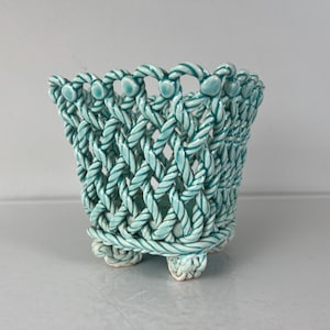 May include: A ceramic planter with a woven design in shades of teal and white. The planter has four small feet and is perfect for displaying small plants or flowers.
