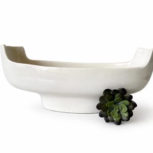 May include: A white ceramic decorative bowl with a boat-like shape, featuring raised ends. A dark green succulent plant is placed next to the bowl. The bowl is designed for decorative purposes.