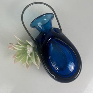 May include: A dark blue glass vase with a teardrop-shaped design and a metal handle. The vase has a small opening at the top and a large, open space in the center. A succulent plant with green and pink leaves is next to the vase.