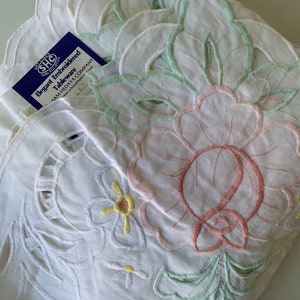 May include: White embroidered tablecloth with a floral design in pink, green, and yellow. The tablecloth has a scalloped edge and the text "Elegant Embroidered Tableware Sam Hedaya Company" is visible on a blue tag.