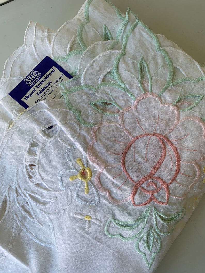 May include: White embroidered tablecloth with a floral design in pink, green, and yellow. The tablecloth has a scalloped edge and the text "Elegant Embroidered Tableware Sam Hedaya Company" is visible on a blue tag.