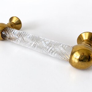 May include: A cabinet handle featuring a clear, twisted glass center bar connected to two polished brass spherical end mounts. The hardware has a cylindrical design with a spiral pattern etched into the glass, finished with metallic gold-toned ends.