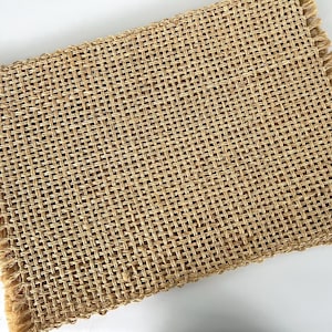 May include: A rectangular placemat made of woven natural jute fibers. The placemat has a fringed edge and a tightly woven pattern. The color is a light tan.