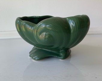 May include: A green ceramic planter shaped like a seashell with a swirl design. The planter has a wide, shallow bowl and a base with three feet.