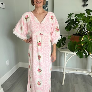 May include: A pink and white patterned maxi dress with a V-neck and short sleeves. The dress features floral embroidery and fringe detailing on the sleeves and hem. The dress is made of a soft, textured fabric.