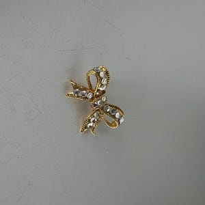 May include: A gold-toned bow brooch with clear, sparkling rhinestones. The bow design features a textured surface and is set against a neutral background. This jewelry item is suitable for adding a touch of elegance to any outfit.