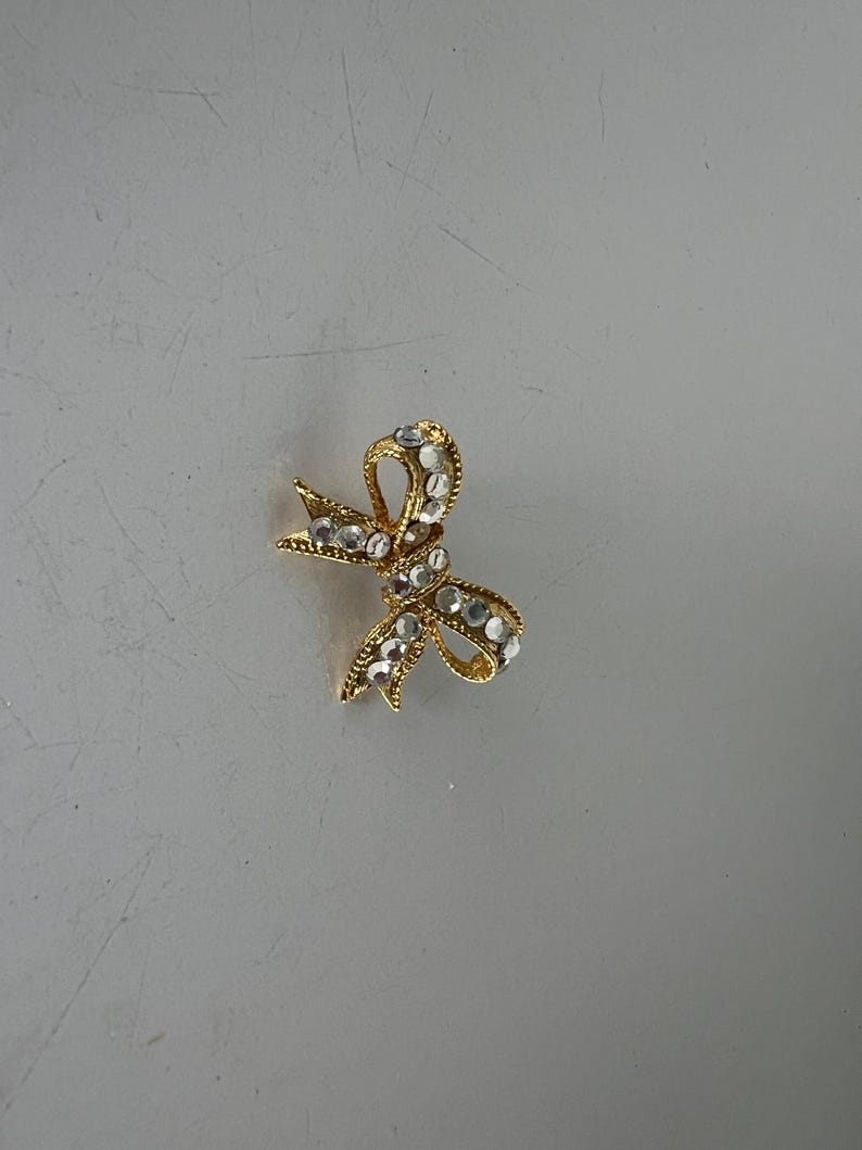 May include: A gold-toned bow brooch with clear, sparkling rhinestones. The bow design features a textured surface and is set against a neutral background. This jewelry item is suitable for adding a touch of elegance to any outfit.