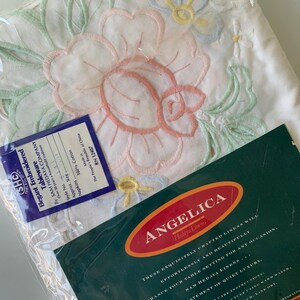 May include: A white embroidered tablecloth featuring a floral design in pink, green, blue, and yellow. The tablecloth is packaged in plastic and has a label that reads "Angelica Hedaya Linens". The label also includes the text "These exquisitely crafted linens will effortlessly and beautifully enhance your table setting for any occasion. A statement of quiet luxury."