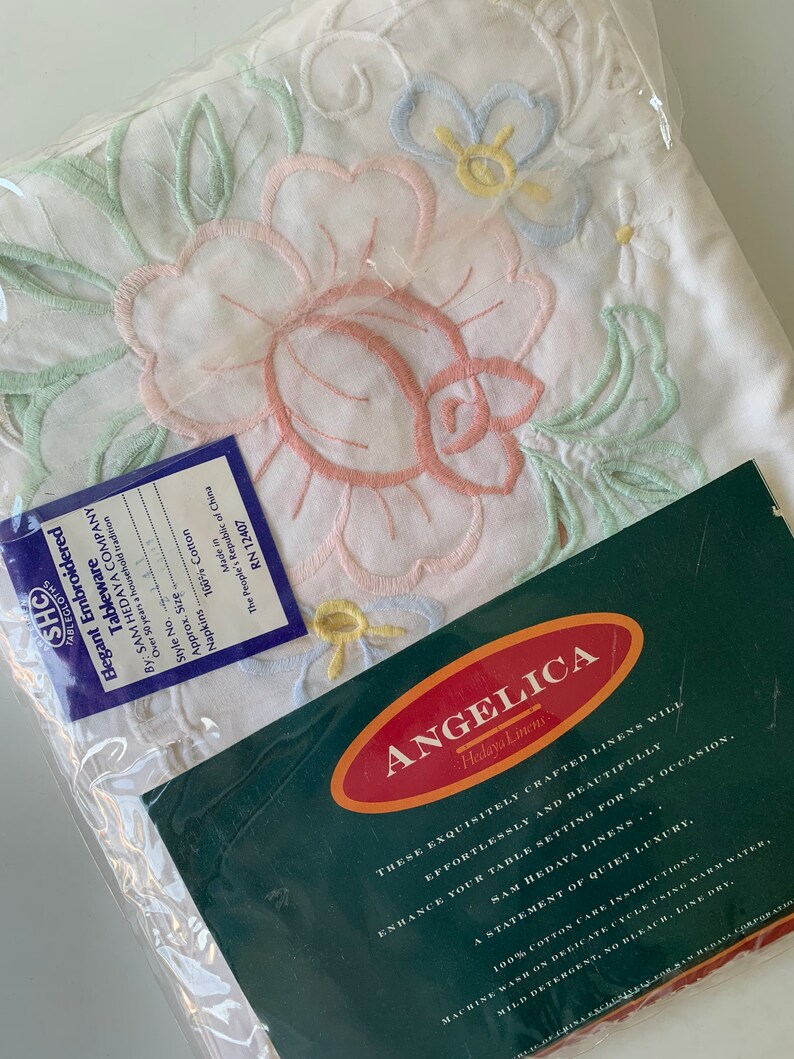 May include: A white embroidered tablecloth featuring a floral design in pink, green, blue, and yellow. The tablecloth is packaged in plastic and has a label that reads "Angelica Hedaya Linens". The label also includes the text "These exquisitely crafted linens will effortlessly and beautifully enhance your table setting for any occasion. A statement of quiet luxury."