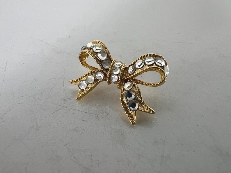May include: A gold-toned bow brooch with clear, sparkling rhinestones. The bow design features a textured surface and is adorned with multiple rhinestones. A classic jewelry accessory.