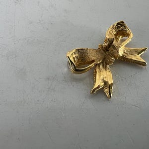 May include: A gold-colored bow-shaped brooch. The brooch has a textured surface and a pin backing. The bow design features detailed folds and pointed ends, suggesting a classic, elegant style. The brooch is likely made of metal.