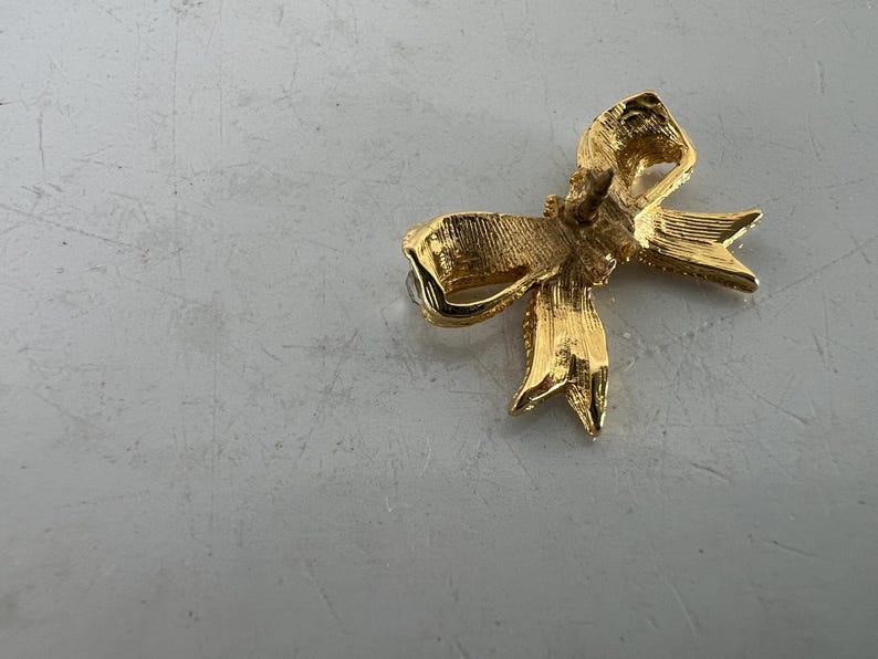 May include: A gold-colored bow-shaped brooch. The brooch has a textured surface and a pin backing. The bow design features detailed folds and pointed ends, suggesting a classic, elegant style. The brooch is likely made of metal.