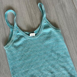Puede incluir: Camiseta sin mangas a rayas turquesa y blancas con tirantes finos. La parte superior tiene un escote redondo y un patr&oacute;n de rayas horizontales. La tela parece ser un material de punto. La camiseta sin mangas se muestra sobre una superficie de madera gris claro.