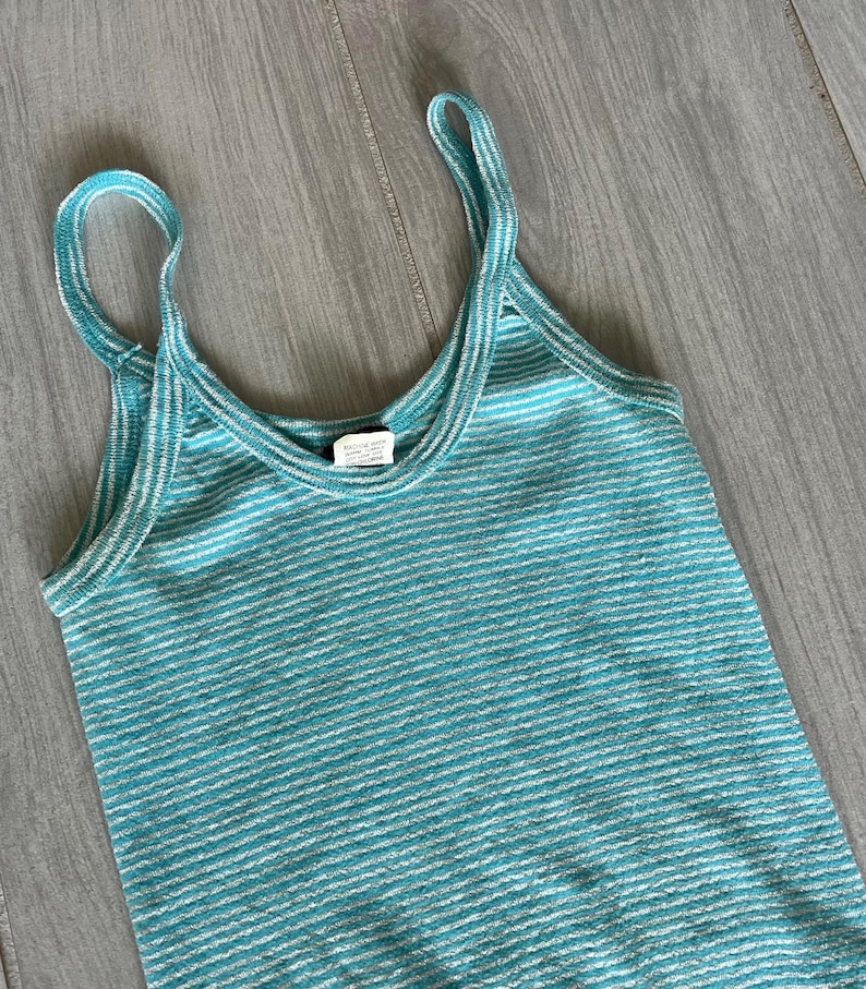 Puede incluir: Camiseta sin mangas a rayas turquesa y blancas con tirantes finos. La parte superior tiene un escote redondo y un patr&oacute;n de rayas horizontales. La tela parece ser un material de punto. La camiseta sin mangas se muestra sobre una superficie de madera gris claro.
