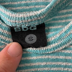 Puede incluir: Primer plano de una camisa a rayas turquesa y blanca. La camisa tiene una textura acanalada y una peque&ntilde;a etiqueta negra con las letras "BDG" y la talla "S" impresas. La tela es suave y parece ser un material de punto.