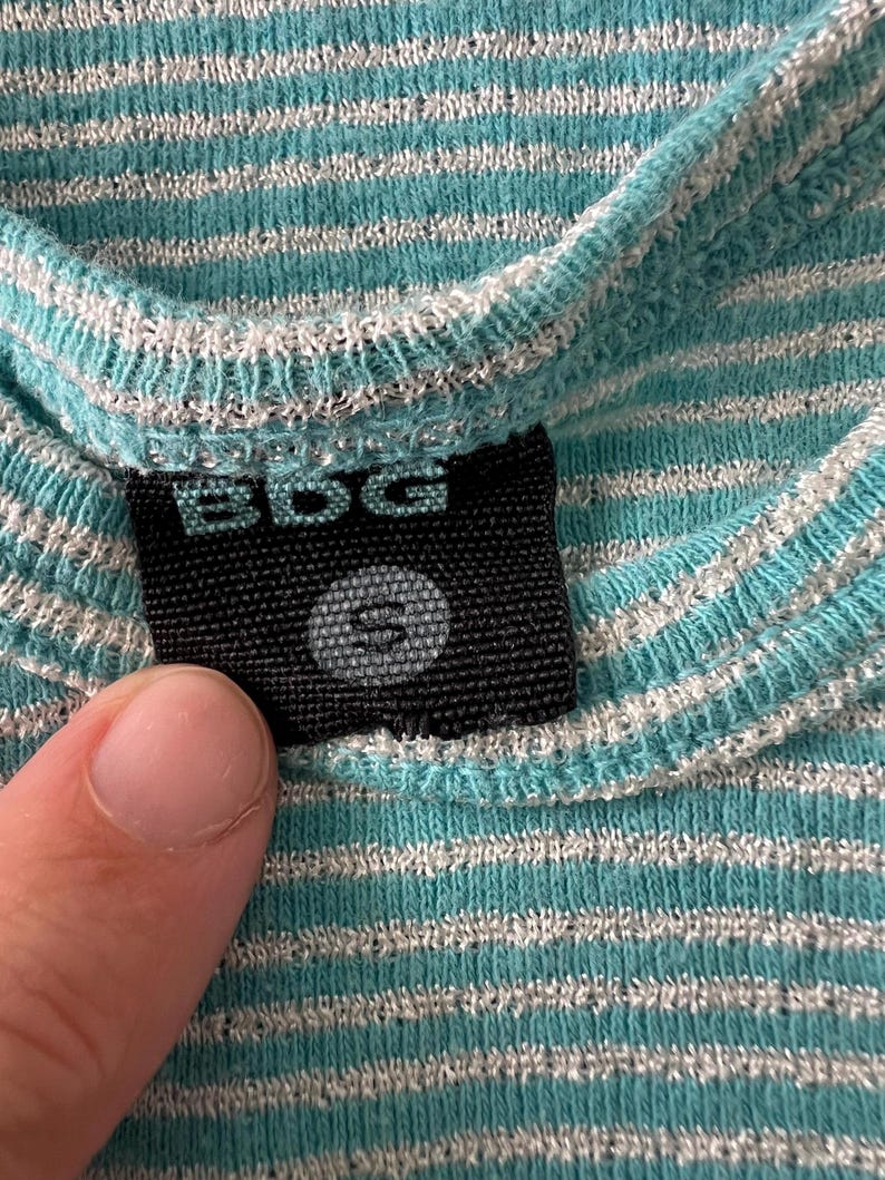 Puede incluir: Primer plano de una camisa a rayas turquesa y blanca. La camisa tiene una textura acanalada y una peque&ntilde;a etiqueta negra con las letras "BDG" y la talla "S" impresas. La tela es suave y parece ser un material de punto.