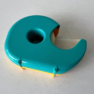 May include: A teal and yellow tape dispenser. The dispenser has a rounded shape with a hole in the center and a blade for cutting tape. The tape roll is visible. The dispenser is made of plastic.