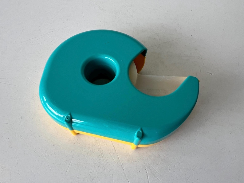 May include: A teal and yellow tape dispenser. The dispenser has a rounded shape with a hole in the center and a blade for cutting tape. The tape roll is visible. The dispenser is made of plastic.