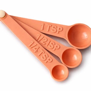 May include: Three peach-colored measuring spoons, labeled 1 TSP, 1/2 TSP, and 1/4 TSP, are fanned out on a white background. The spoons are made of plastic and are connected at the top with a small, round, off-white piece.