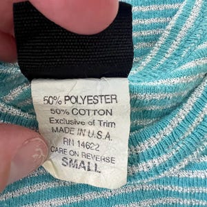 Puede incluir: Primer plano de una etiqueta de ropa con el texto "50% POLYESTER, 50% COTTON, MADE IN U.S.A., SMALL." La etiqueta se sostiene contra una tela a rayas azules y blancas. Tambi&eacute;n se ve el texto "CARE ON REVERSE."