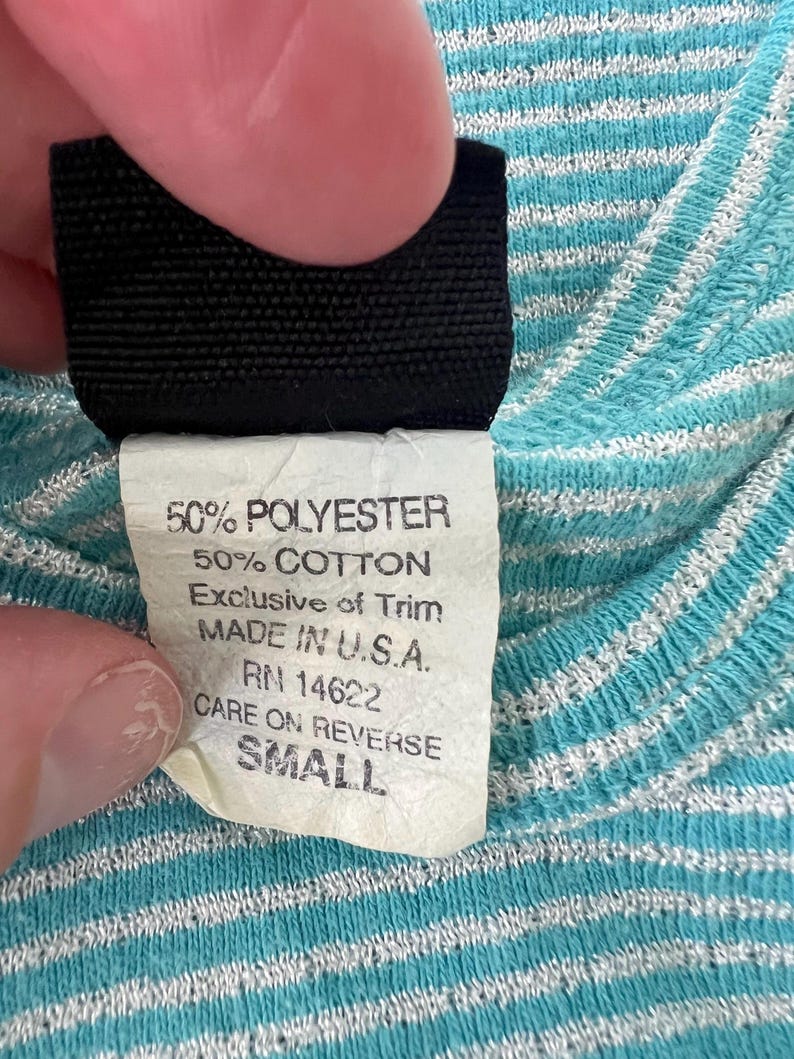 Puede incluir: Primer plano de una etiqueta de ropa con el texto "50% POLYESTER, 50% COTTON, MADE IN U.S.A., SMALL." La etiqueta se sostiene contra una tela a rayas azules y blancas. Tambi&eacute;n se ve el texto "CARE ON REVERSE."