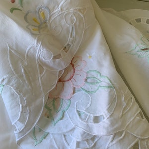 May include: White cotton fabric with delicate embroidered floral designs in pastel colours. The fabric features intricate cutouts and scalloped edges.