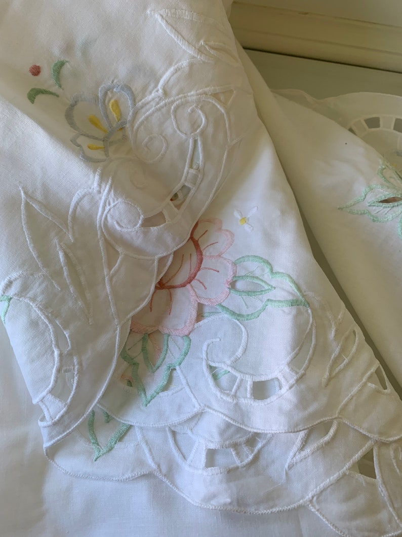 May include: White cotton fabric with delicate embroidered floral designs in pastel colours. The fabric features intricate cutouts and scalloped edges.