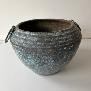May include: A weathered, dark gray metal bowl with a textured surface and two ring handles. The bowl has a wide opening and a rounded base. The exterior has a mottled, blue-green patina, suggesting age and use. A decorative piece.