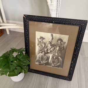 May include: A black-and-white vintage photograph in a textured black frame, depicting a group of seven individuals in costumes, including hats and props, arranged in a seated and standing pose. The image is set against a tan matting.