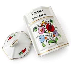 May include: A white ceramic spice container with a matching lid, featuring gold trim and a colorful floral design. The text "Paprika" and "Süß - Sweet" is printed in black on the front. The lid is decorated with small red chili pepper illustrations.