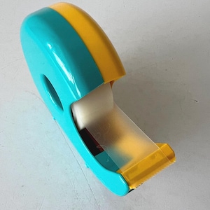 May include: A teal and yellow tape dispenser with a clear tape roll. The dispenser has a rounded top and a serrated edge for cutting the tape. The tape dispenser is made of plastic.