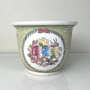 May include: A small, round ceramic planter with a floral design. The planter features a light green background with a white rim and a central oval design with pink, blue, and yellow flowers. The design is framed by a green wreath.