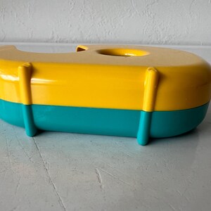 May include: A colorful tape dispenser in a boat shape. The top is yellow, the bottom is teal, and it has yellow vertical supports. The dispenser has a circular opening on top for the tape roll. A fun and functional desk accessory.