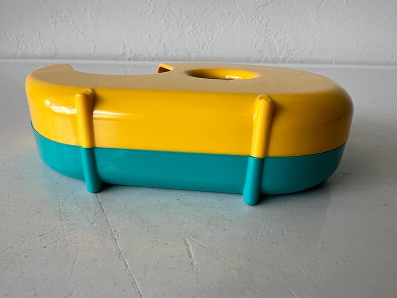 May include: A colorful tape dispenser in a boat shape. The top is yellow, the bottom is teal, and it has yellow vertical supports. The dispenser has a circular opening on top for the tape roll. A fun and functional desk accessory.