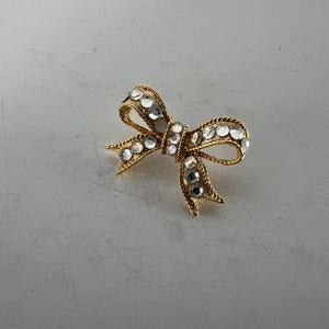 May include: A gold-toned bow brooch adorned with clear, sparkling rhinestones. The bow design features intricate detailing and a classic shape, suitable for adding a touch of elegance to any outfit. The brooch is a timeless accessory.