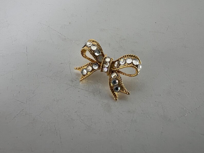 May include: A gold-toned bow brooch adorned with clear, sparkling rhinestones. The bow design features intricate detailing and a classic shape, suitable for adding a touch of elegance to any outfit. The brooch is a timeless accessory.